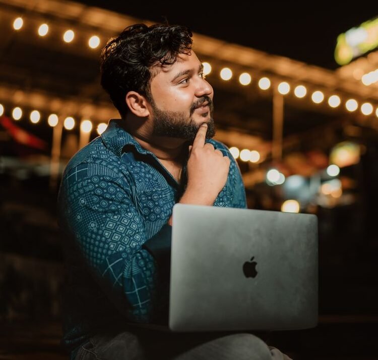 A thoughtful adult man with a beard using a laptop at night in a brightly lit outdoor setting.