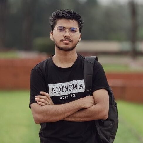 Young man with glasses in casual attire, standing confidently in a garden setting.