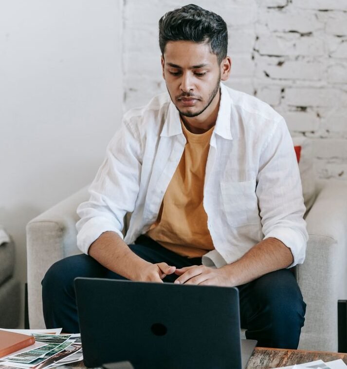 Creative young Hispanic male photographer sitting at table with photo camera and printed photos and working on laptop in modern loft style workplace at home